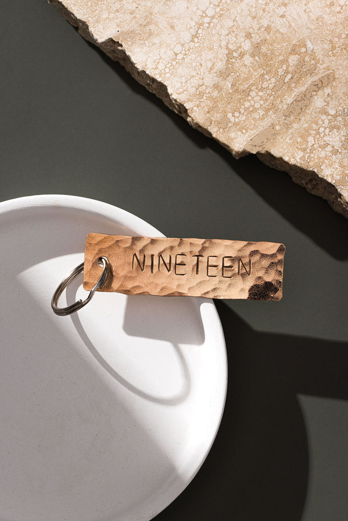 A keyring made from Bronze that has been planished and engraved with the word NINETEEN. The keyring is laid upon a small, white dish which is also on top of a moss green background. There is a partial slab of rugged, white marble in the top right hand corner of the frame. The bronze keyring is made by Empire Copper.