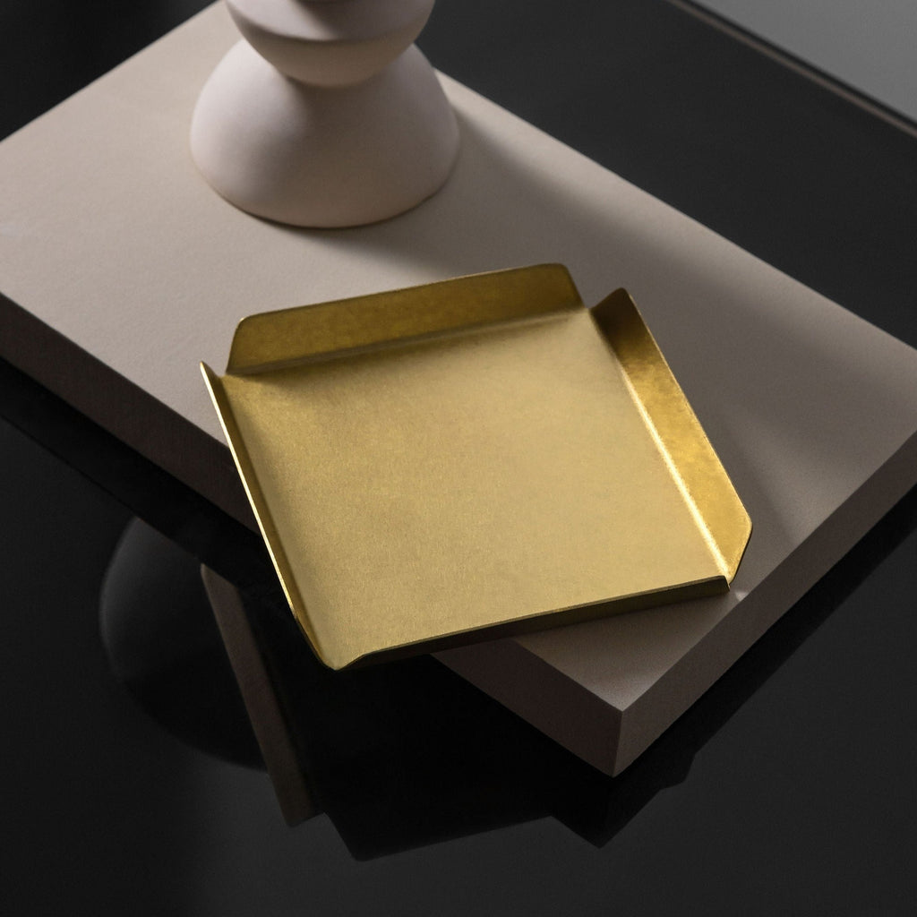 Pictured is a square 100mm X 100mm real brass tray with raised, bevelled edges to each side. The tray is sitting upon a white block, with the base of a white, thin vase pictured in the background. The white vase, white block and brass tray are upon a black, reflective table top. The brass tray is brushed and matte and is made by Empire Copper.