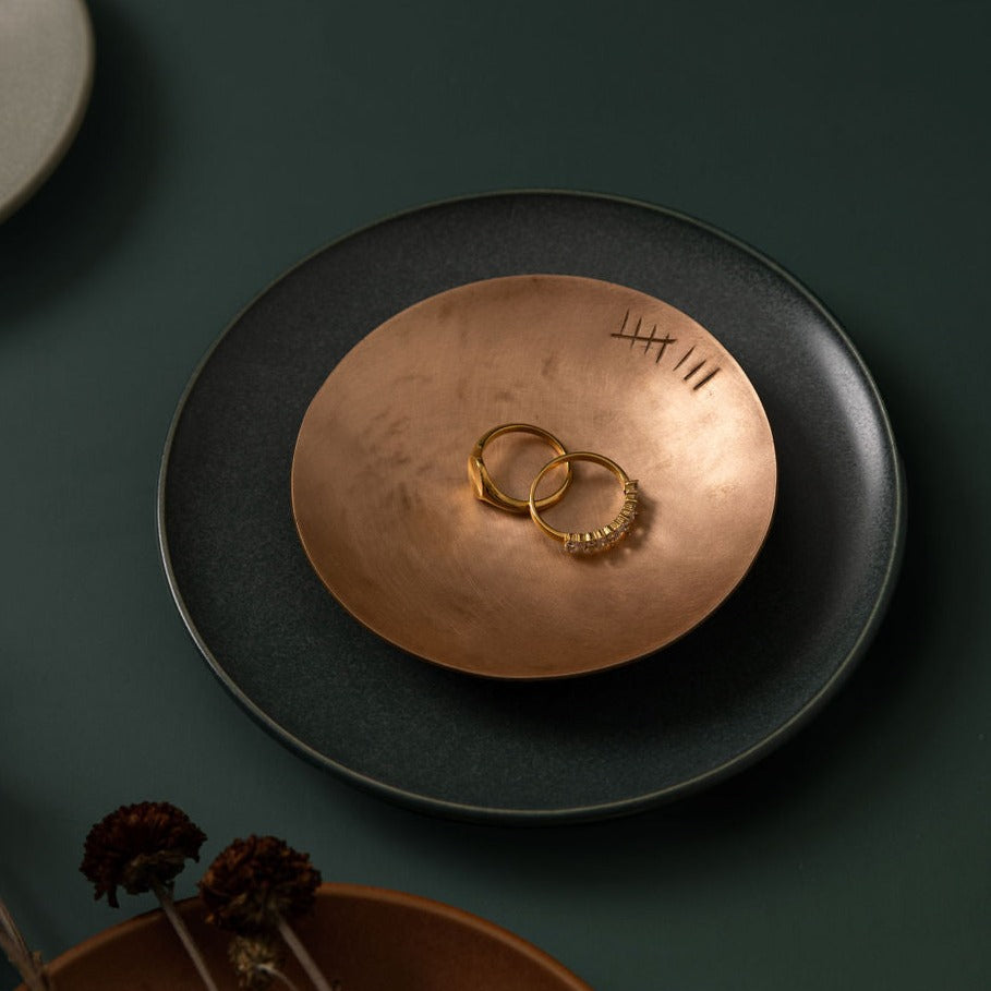 A small, round, bronze trinket dish with the tally number eight engraved on the right hand side just inside the rim. There is two gold rings inside the trinket dish. The trinket dish is sat upon a dark green saucer and the saucer is upon an emerald green background. The trinket dish is made by Empire Copper.