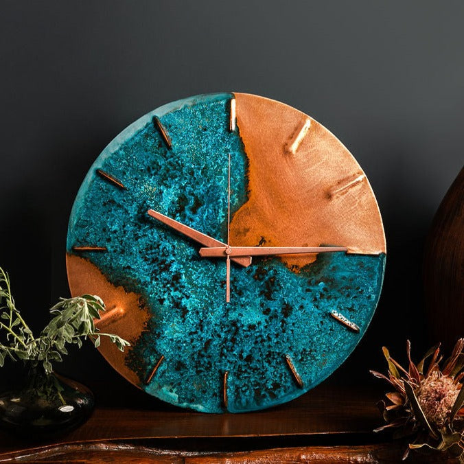 A copper wall clock with a vibrant, blue patina sits atop a wooden bench and in front of navy a background. The clock is made by the company Copper Brass and Bronze and Empire Copper by  Hayes Home.