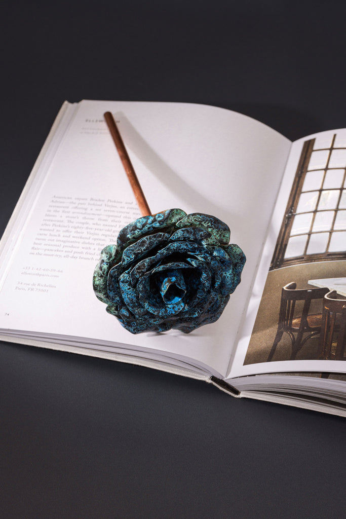 A rose made with a copper stem and a blue patina, copper rose head; lays flat across an open book. The open book is placed upon a dark navy surface. The copper rose is made by Empire Copper.