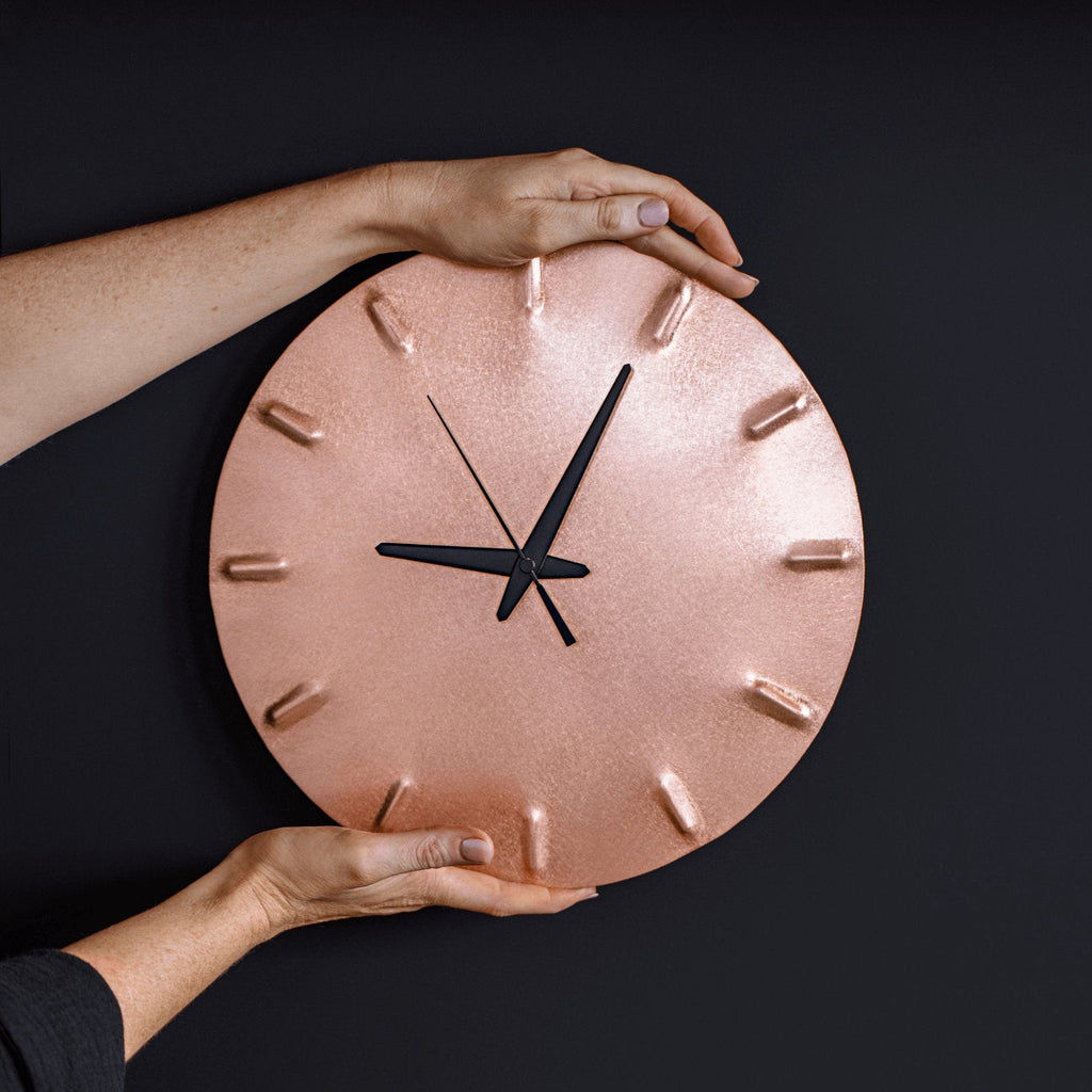 A round, brushed copper clock with black hands point to five minutes past nine. Two hands grasp the top and bottom of the clock, holding it against a navy wall.