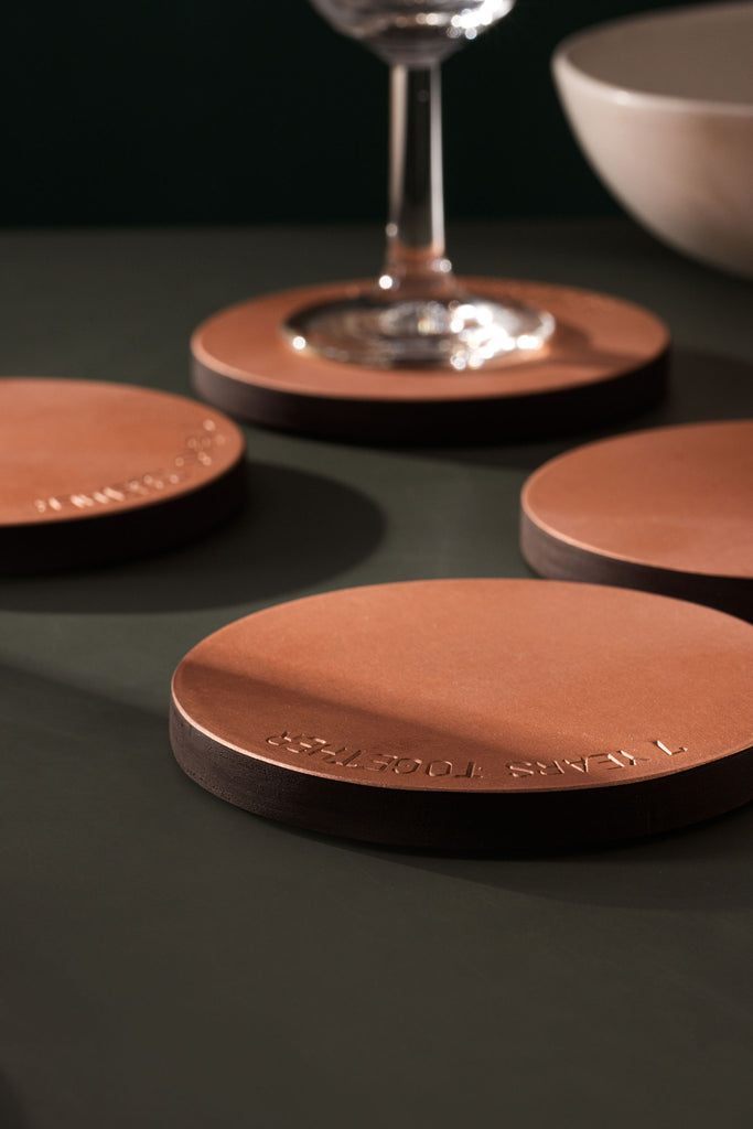A portrait shaped photo with four, round copper coasters placed upon an olive coloured table top. The coasters are copper and wood. They coasters are engraved "7 YEARS TOGETHER". The coaster at the back has a wine glass placed upon it. The coasters are made by Empire Copper.