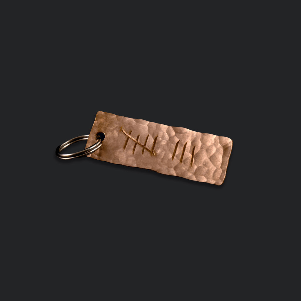 A keyring made from planished bronze and engraved with the eight tally. The keyring is on a navy background. The keyring is made by Empire Copper.