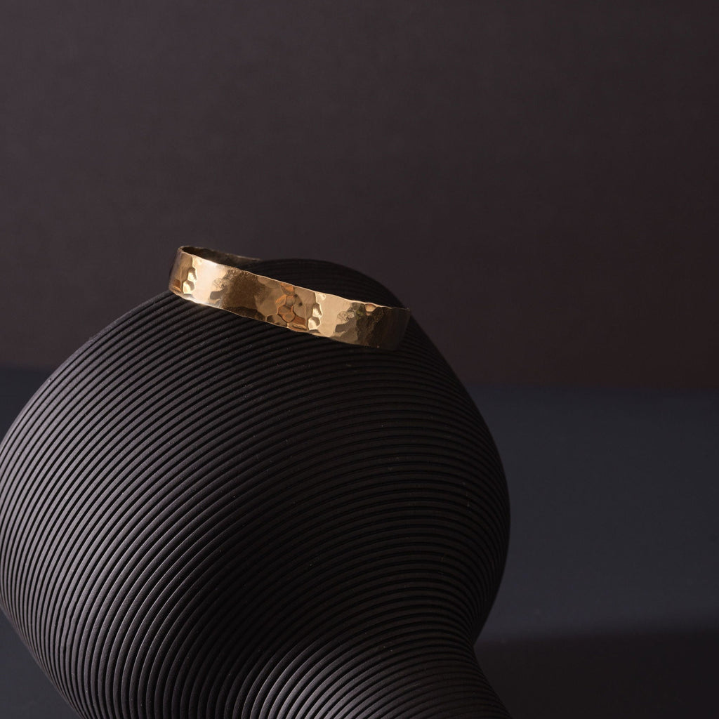 A planished brass cuff, twinkling in the light. The cuff is placed upon a black, turned over vase. The vase is upon a dark navy tabletop and the background is charcoal in colour. The brass cuff bracelet is made by Empire Copper.