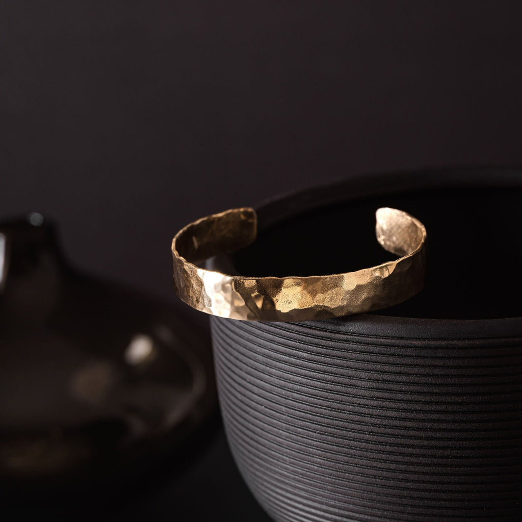 A planished brass cuff, sparkling where the light is hitting it. The brass cuff placed is resting upon a black vase with horizontal ribbed detail. The vase is upon a dark navy tabletop and the background is charcoal in colour. The brass cuff bracelet is made by Empire Copper.
