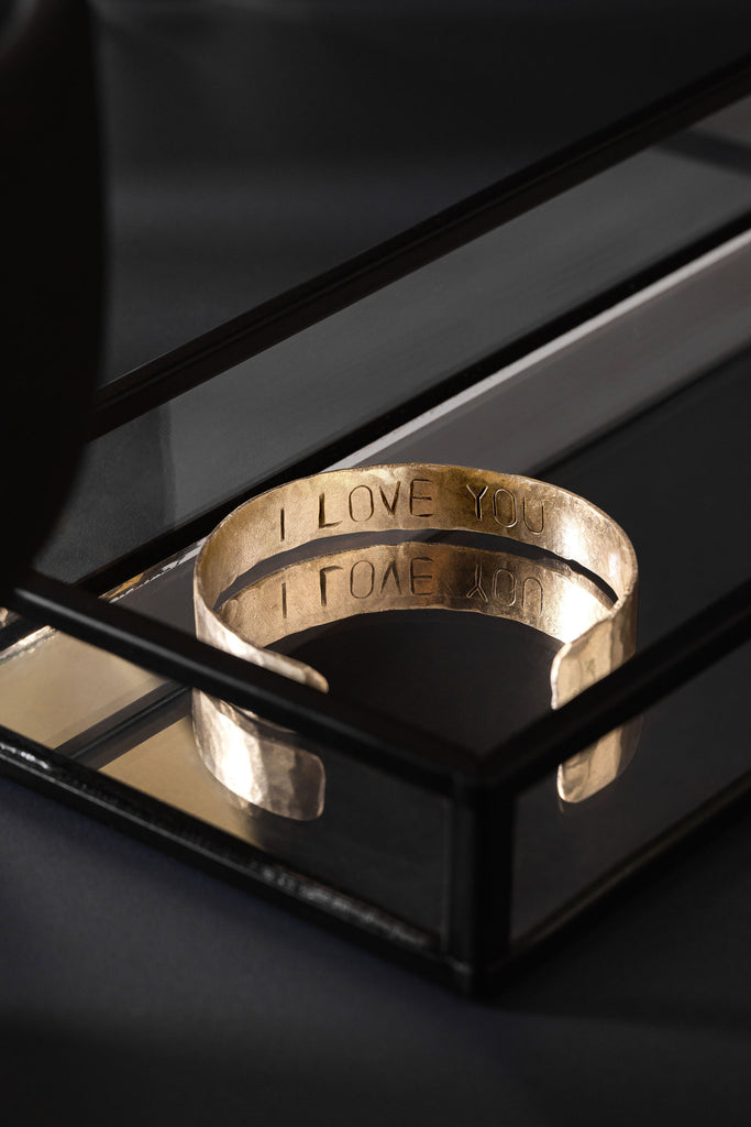 A brass cuff bracelet, photographed upon a glass tray with a mirror base. The cuff is inverted and there is engraving on the inside that reads "I LOVE YOU". The tray the cuff bracelet is in, is placed upon a charcoal coloured tabletop. The brass cuff bracelet is made by Empire Copper.