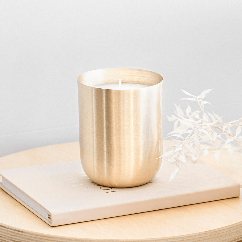 A light wooden table with a small vanilla coloured book on top. A large brass candle vessel sits atop the book. To the right of the brass vessel is a small bunch of white, whimsical leaves. The brass candle vessel is made by Empire Copper.