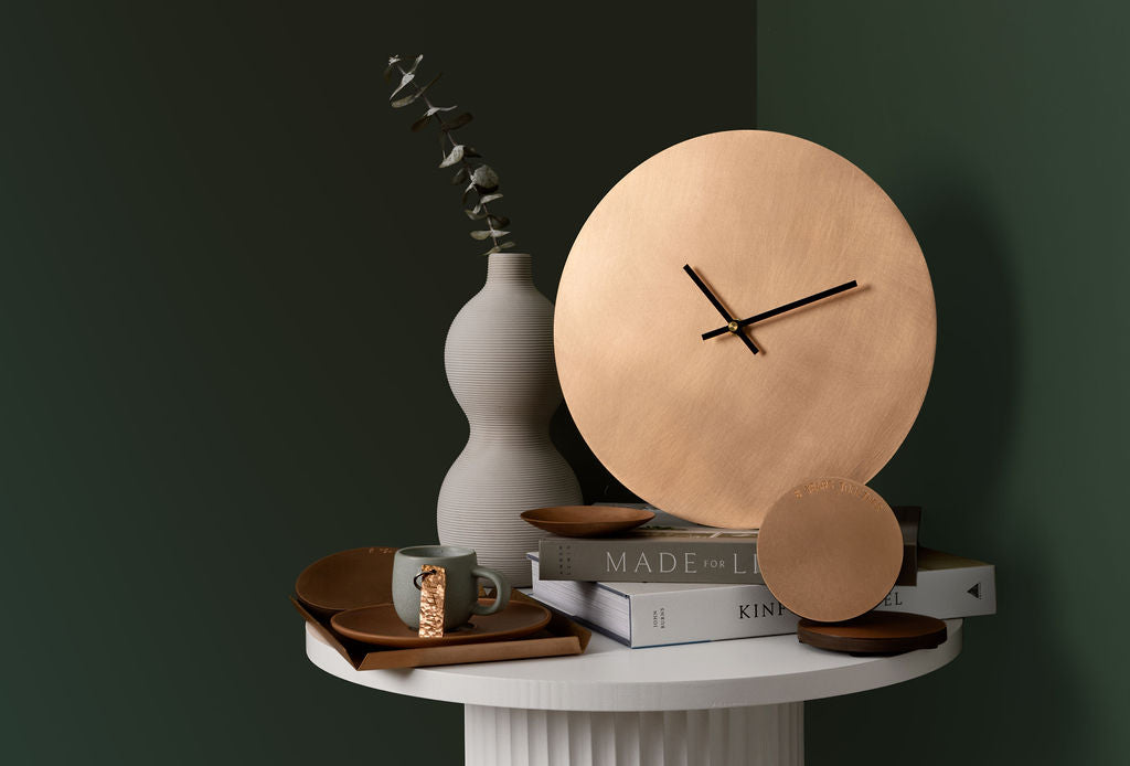 An olive green background with a white round table. On top of the table is a round clock, round coaster, trinkey tray, key ring and rectangle tray all made from bronze. Next to the clock is a tall white wave-like vase; products made by Empire Copper.