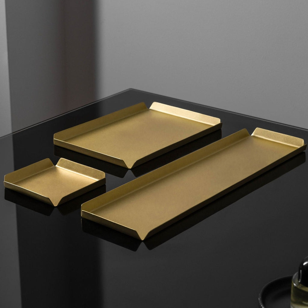 Pictured is a square 100mm X 100mm, a rectangle 235mm x  150mm and a long 100mm x 400mm real brass tray with raised, bevelled edges to each side. The trays are photographed atop a black, reflective surface. The brass trays are finished with a brushed and matte look and are made by Empire Copper.