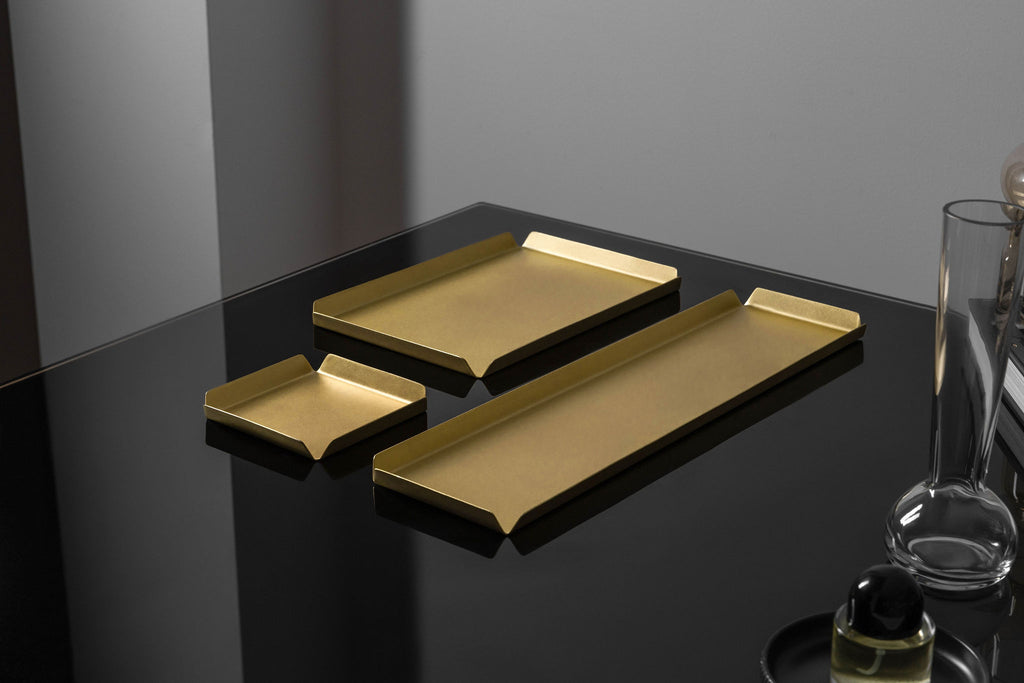 A small, square shaped brass tray, behind that is a medium shaped brass tray and to the right of both of those trays is a long, rectangle shaped brass tray. All the trays have raised edges. The trays are photographed upon a black reflective surface. The brass trays are made by Empire Copper.