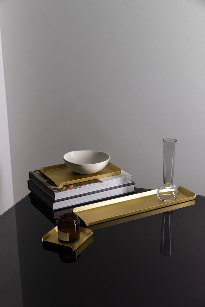 A small, square brass tray, with an amber jar candle on top, a long, rectangular brass tray with a thin glass vase placed on one side and a stack of books with a medium, rectangular brass tray on top. There is a white bowl on top of the medium brass tray. The items are styled upon a black, reflective surface. The background wall is white. The trays are made by Empire Copper.