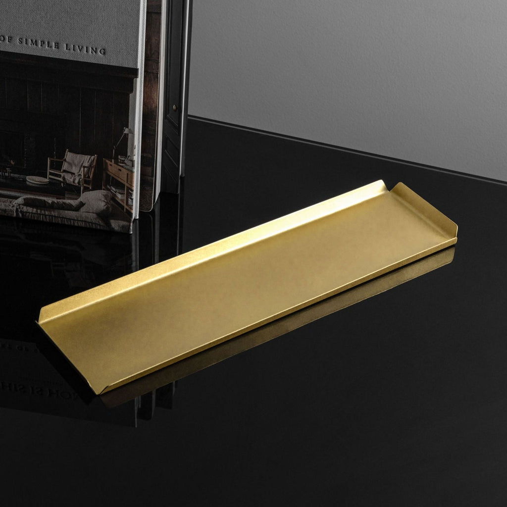 A long, rectangle shaped brass tray with raised edges upon a black reflective surface. The background wall is white and to the left of the tray is the coffee table book This Is Home: The Art of Simple Living. The brass tray is made by Empire Copper.