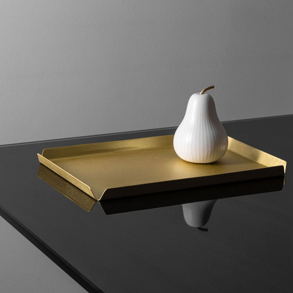 A rectangle shaped brass tray with the dimensions 235mm W x 150mm D sitting atop a black, reflective surface. Upon the brass tray is a white, ornamental pear shaped object. The background of the image is right. The tray is made by Empire Copper.