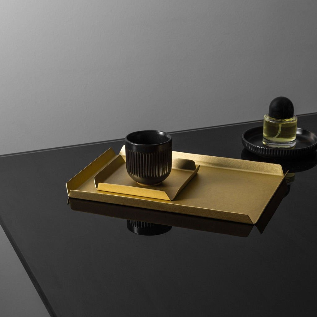 A rectangle shaped brass tray with the dimensions 235mm W x 150mm D with a small brass square tray with the dimensions 100mm x 100mm and a small, black vase sitting atop. To the back right of the tray is a black dish with a perfume bottle on top. All the items are styled upon a black, reflective surface. The background of the image is right. The tray is made by Empire Copper.