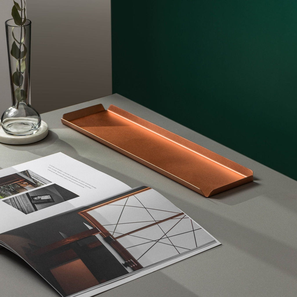 A long, rectangular copper tray that is 400mm W and 100mm D. The tray is upon a white tabletop with an open coffee table book in front. To the left of the tray and book is a clear, glass vase upon a white coaster. The background is emerald green. The copper tray is made by Empire Copper.