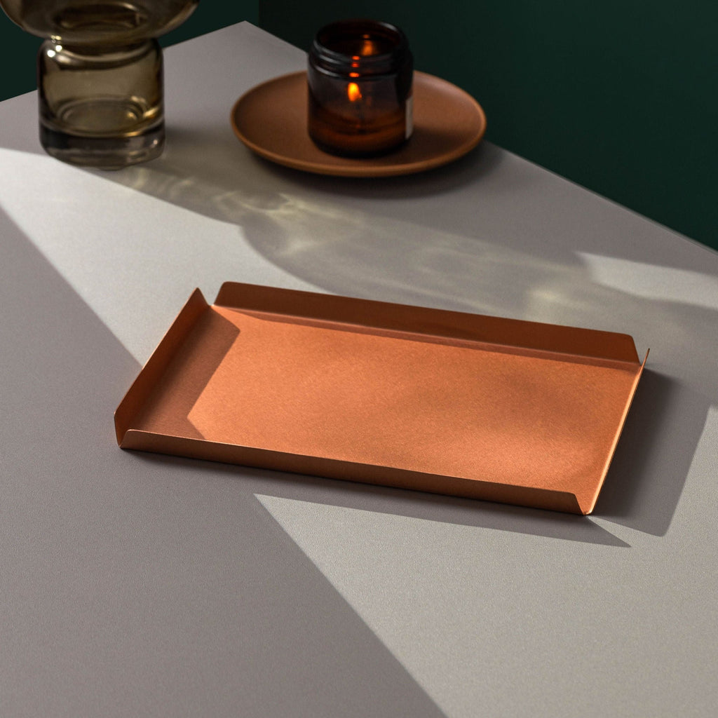 A 235mm W x 150mm D rectangular copper tray with raised edges on all sides upon a white tabletop. The image background in emerald green. Behind the tray is an amber candle burning atop a brown plate and to the left of that is the base of a vase half in the image. The copper tray is made by Empire Copper.