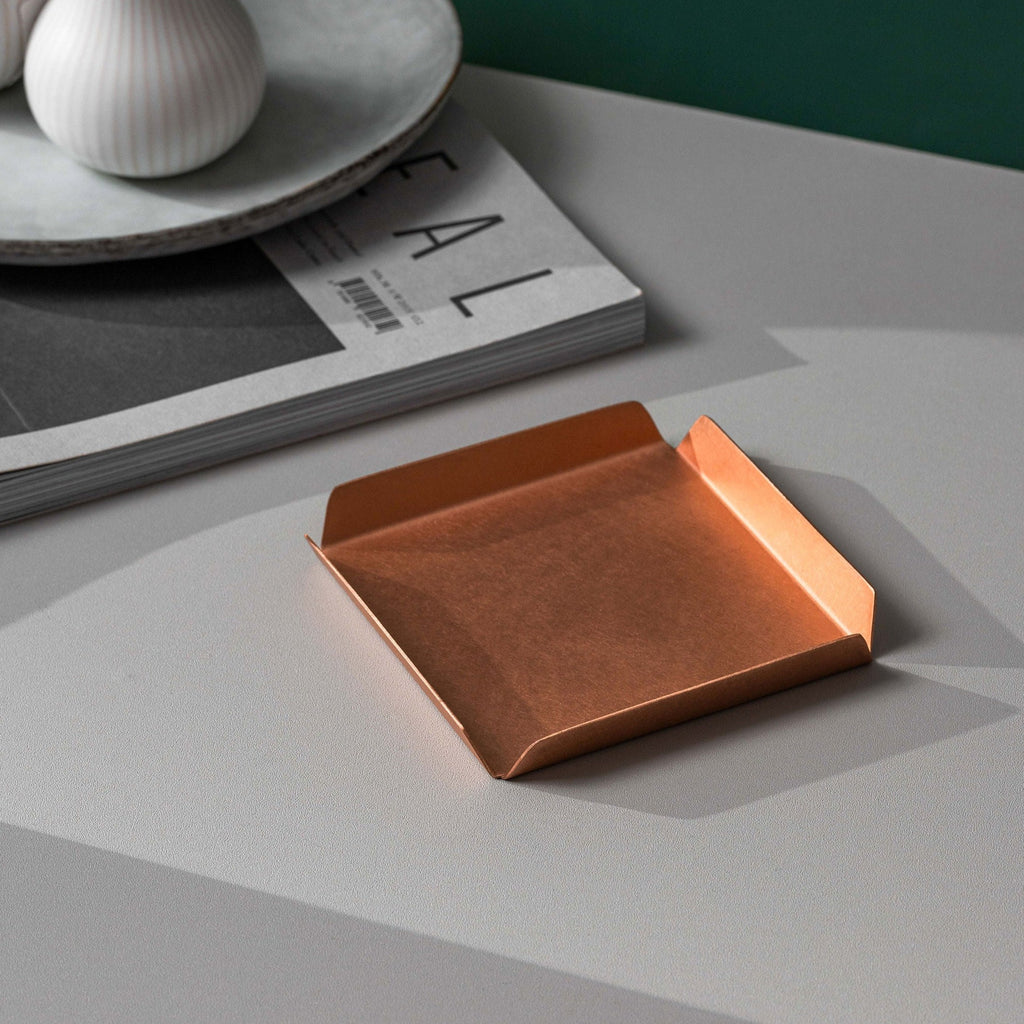 A small square 100mm x 100mm copper tray with raised edges on all sides. The tray is placed upon a white surface with a magazine and white dish in the top left hand corner of the image. The background wall is emerald green. The copper tray is made by Empire Copper.