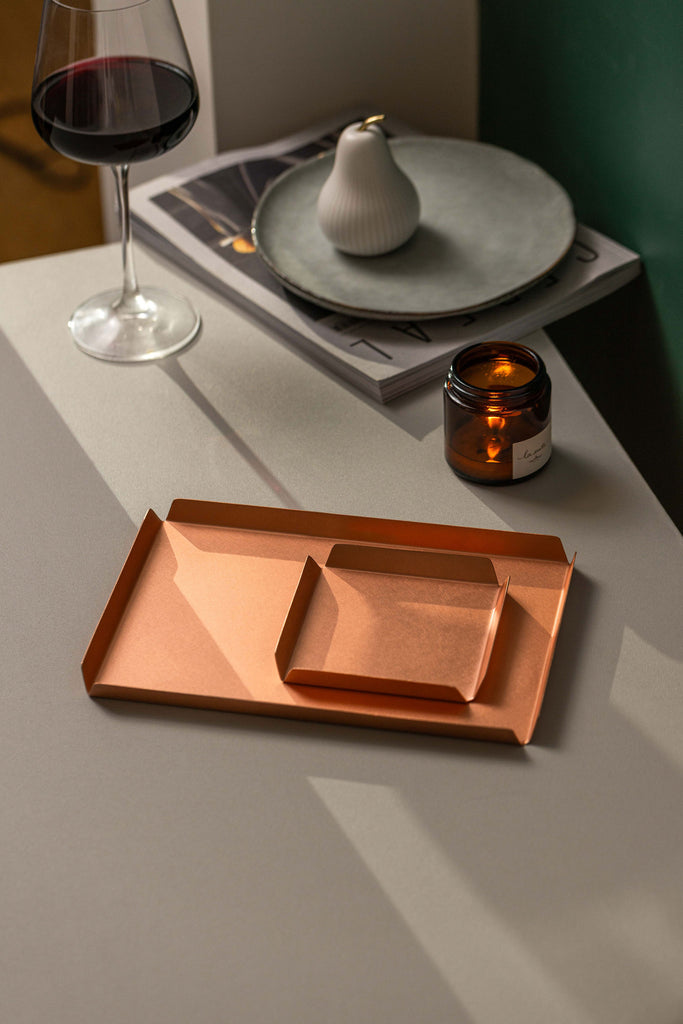 A small square 100mm x 100mm copper tray with raised edges sitting upon a 235mm W x 150mm D copper tray. The trays are placed upon a white surface with a magazine and white dish in the top left hand corner of the image. There is also a glass of red wine to the back left of the trays and a burning candle in an amber vessel behind the trays.The background wall is emerald green. The copper tray is made by Empire Copper.