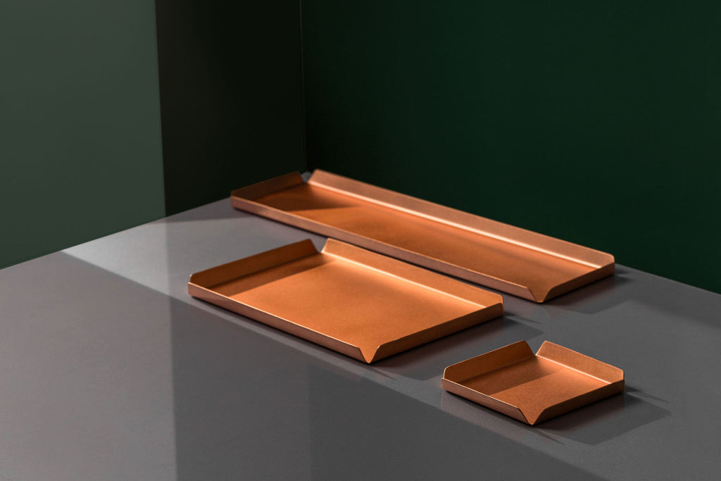 A 100mm x 100mm square copper tray, a 235mm W x 150mm D copper tray and a 400mm W x 100mm D copper tray all sitting upon a white surface. The background of the image is emerald green. The copper trays are made by Empire Copper.