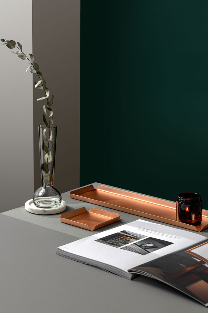 A long, rectangular copper tray that is 400mm W and 100mm D that has a burning candle in an amber jar placed on the right end of the tray. The tray is upon a white tabletop with an open coffee table book in front. To the left of the tray and book is a clear, glass vase upon a white coaster. In front of the tray is a smaller, square copper tray that is 100mm x 100mm. The background is emerald green. The copper trays are made by Empire Copper.