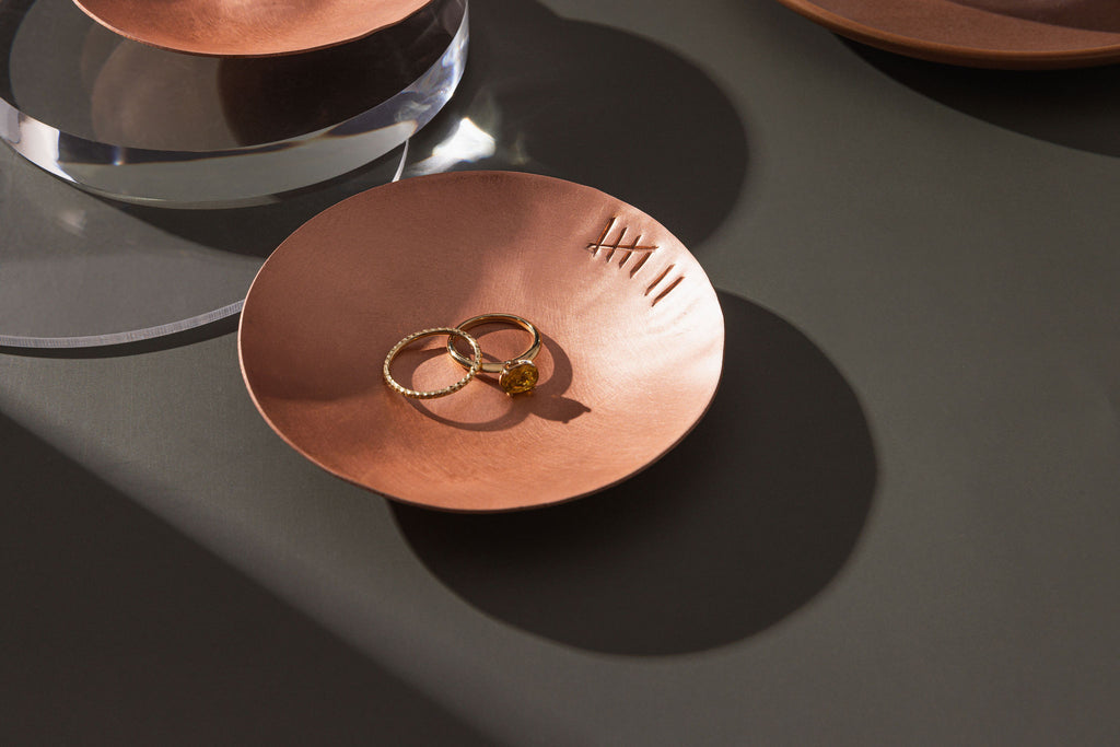 A small, round, matte copper trinket dish with an seven tally engraved along the rim. The dish has been photographed atop a moss green background. Within in the trinket dish there is two gold rings, one with a large amber stone. The trinket dish is made by Empire Copper.