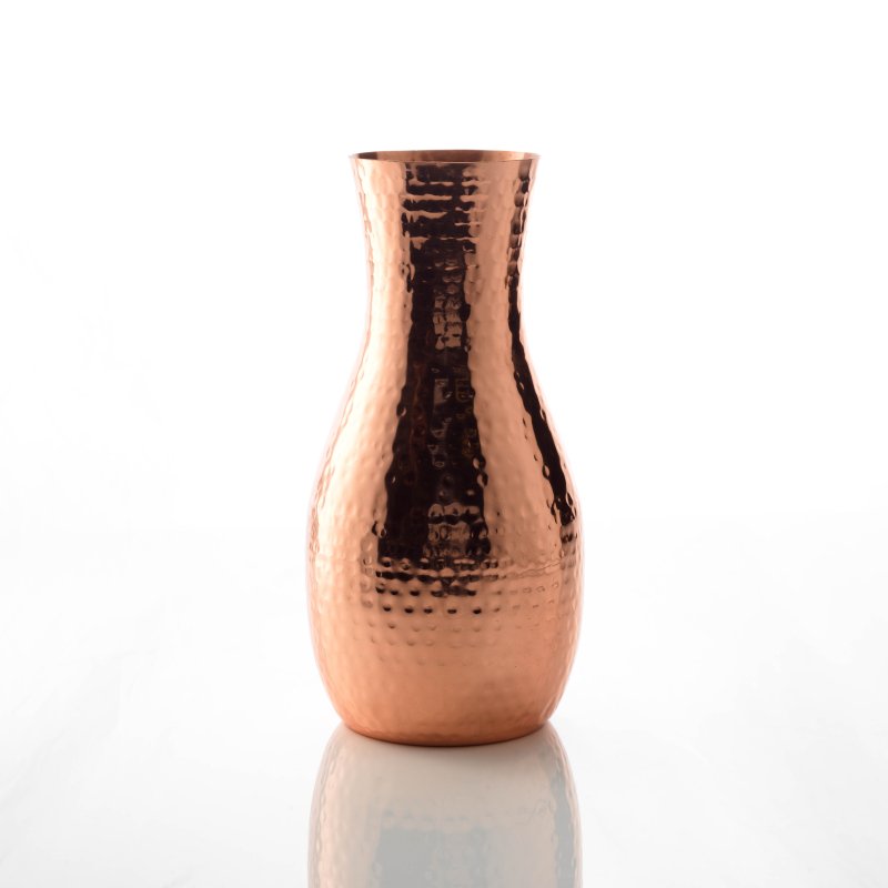 A hammered copper carafe sitting on a white, reflective background. The carafe is sold by Empire Copper by Hayes Home.