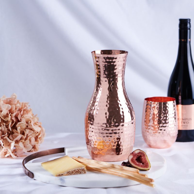 A hammered copper carafe sitting atop a marble cheese board. To the right of the carafe is hammered copper stemless wine glass. The carafe is sold by Empire Copper by Hayes Home.