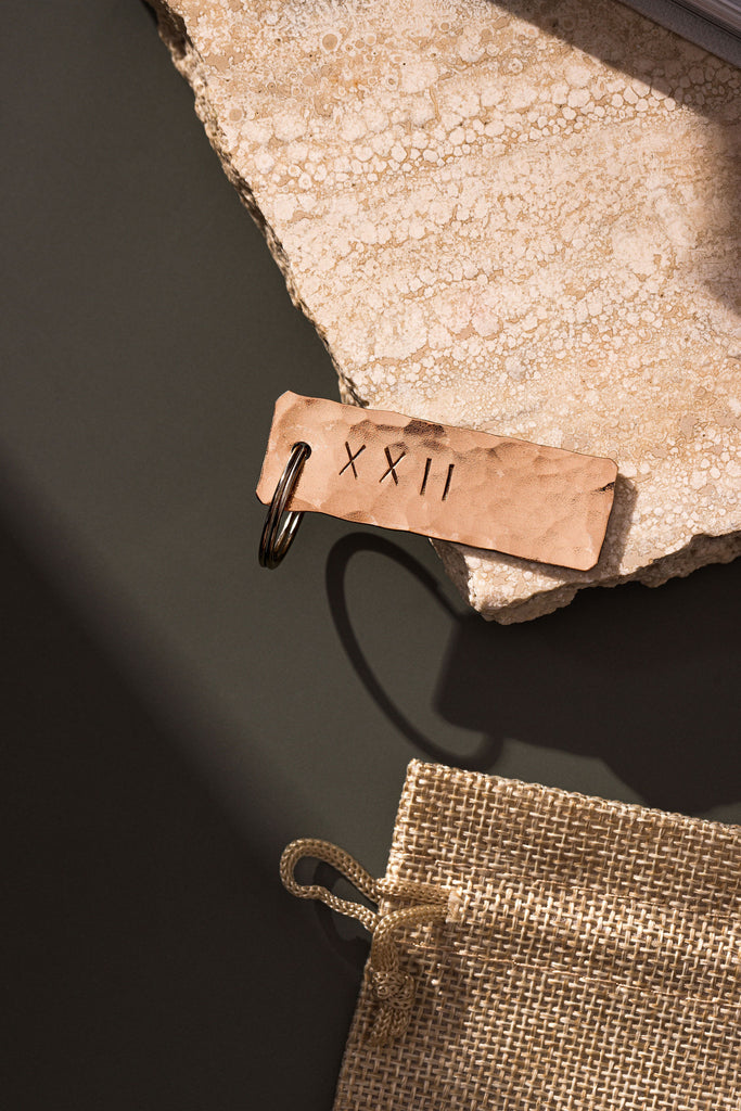 A copper keyring that has been planished and engraved with the roman numeral symbols XXII which translates to the number 22 sitting atop a rugged piece of travertine. The travertine slab is laid upon a moss green background. The copper keyring is made by Empire Copper.