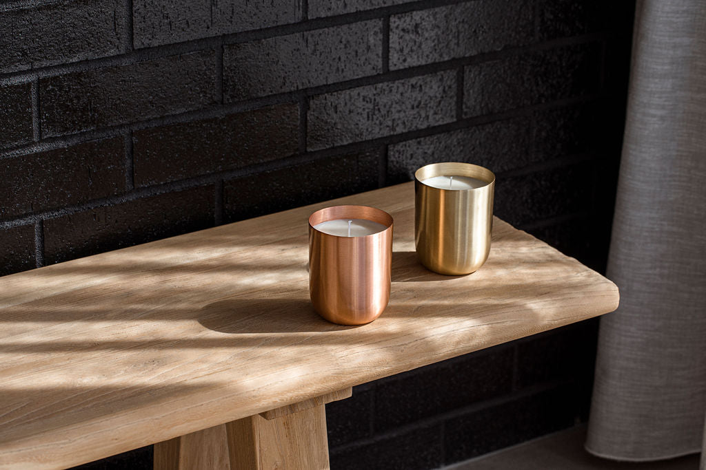 A copper candle vessel and a brass candle vessel on the end of a long wooden bench. The bench is against a black brick wall with a grey curtain to the right, slightly in the image. The candles are made by in Perth by Empire Copper.