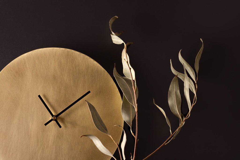 A golden, brass clock with thin black hands sits in the bottom left of the frame, taking up about 50% of the image. Behind the clock is a charcoal coloured wall.  In front of the clock is a thin branch of olive leaves. The image is landscape and clock is made by Empire Copper.