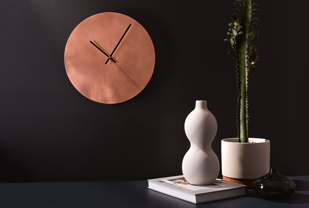 A round, glowing copper clock with thin black hands. The clock is affixed to a dark, charcoal background. To the right of the clock is a long, green cactus that is nestled in a small grey pot. The clock is made by Empire Copper.
