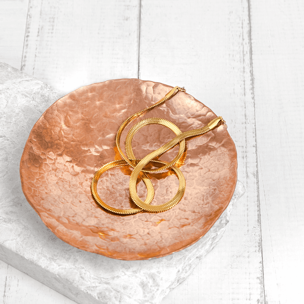 A white wooden background, with a small slab of white marble. On top of the marble slab is a planished copper trinket dish, glistening in the light. Within the copper trinket dish is a thick gold chain. The trinket dish is made by Empire Copper.