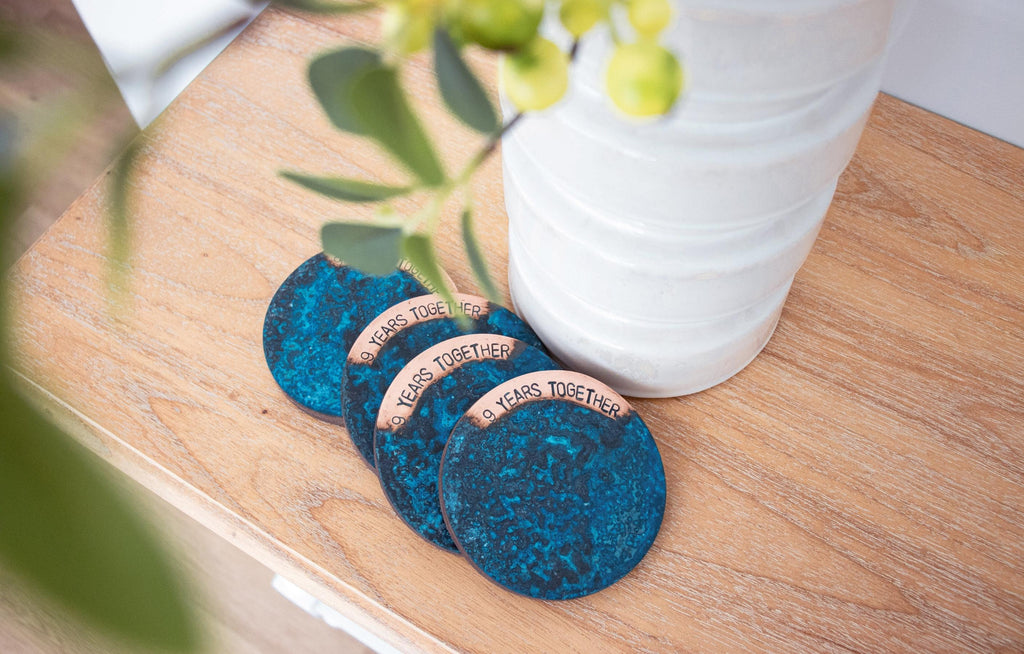 A Set of 4, Blue Patina Coasters engraved with '9 Years Together'. The coaster set is made in Perth by Empire Copper and is pictured sitting atop a light oak, wooden buffet and placed next to a tall white vase.