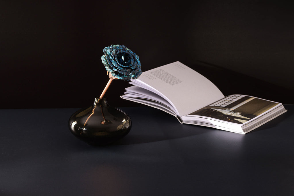 A rose made with a copper stem and a blue patina, copper head sits alone inside a small, glass vase. Behind the rose to the right, lies an open book. The vase, book and rose are all sitting a top a dark, navy table. The copper rose is made by Empire Copper.