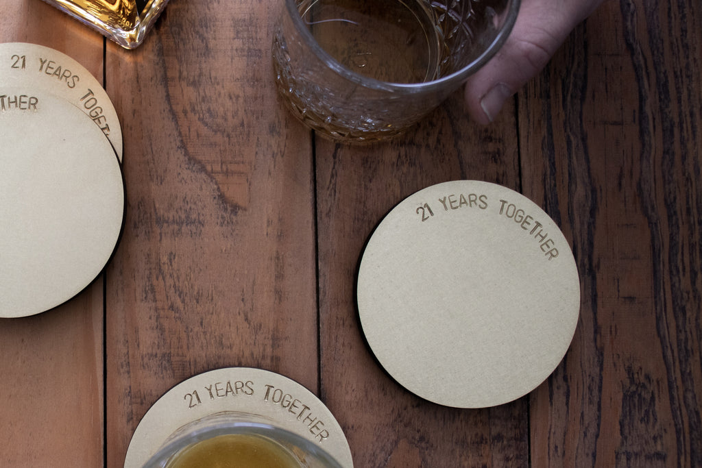 A wooden table with a set of four, brushed brass coasters scattered on top. The coasters are engraved with the words "21 Years Together". The coasters are made by Empire Copper.