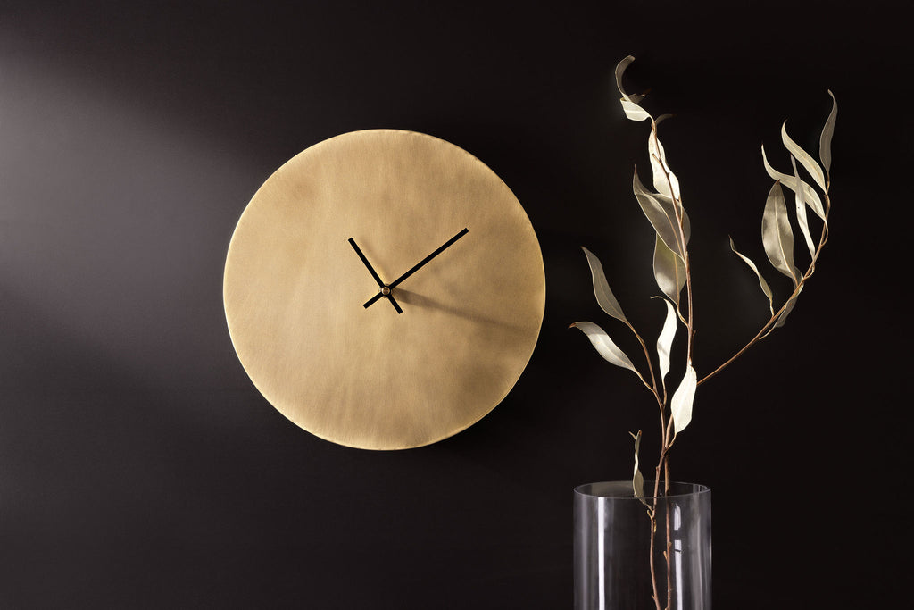 A golden, brass clock with thin black hands affixed to a dark, charcoal coloured wall. To the right of the clock is a glass vase with some long, green leaves. The picture is landscape and clock is made by Empire Copper.