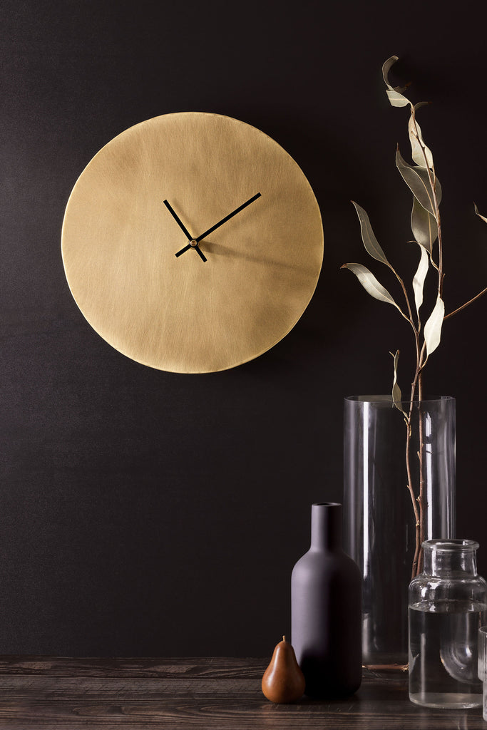 A golden, brass clock with thin black hands affixed to a dark, charcoal coloured wall. To the right of the clock is a glass vase with some long, green leaves. At the base of the vase on a wooden bench is a charcoal coloured vase, a clear vase with water and a brown, small pear shaped ornament. The picture is portrait and clock is made by Empire Copper.
