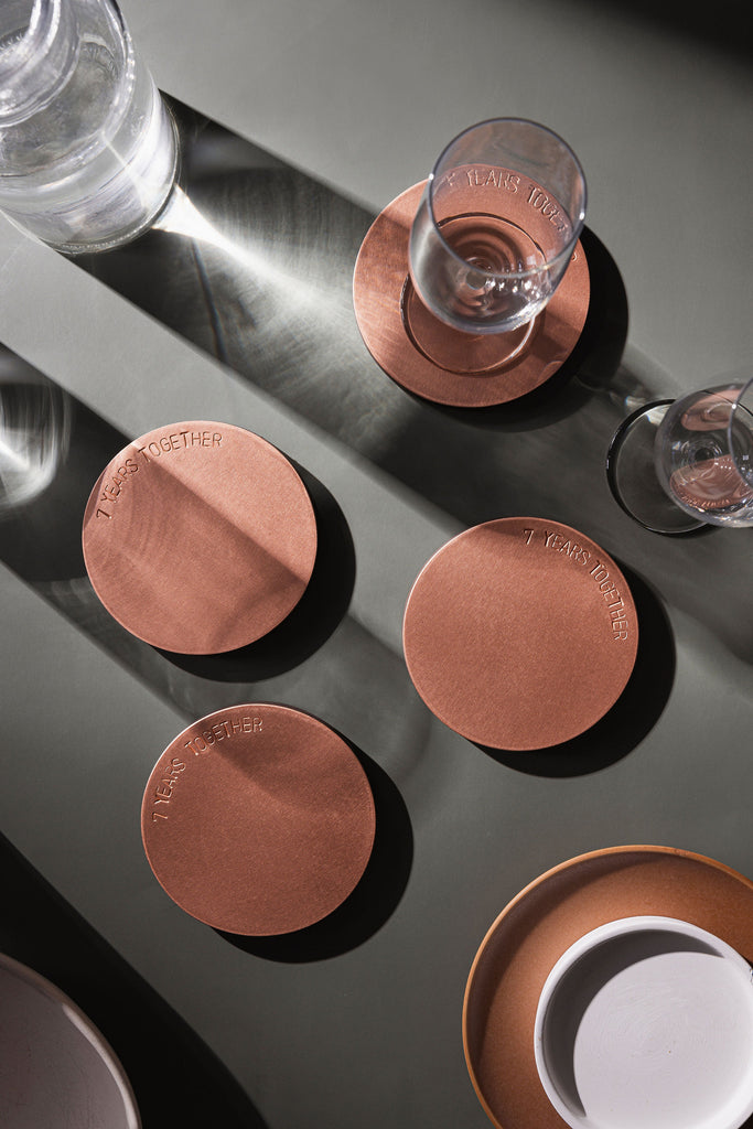 A set of four, copper coasters laying atop a moss green surface. There is a champagne flute atop one of the coasters and shadows draping across the image. This photo is taken as a birdesye view. The coasters are made by Empire Copper.