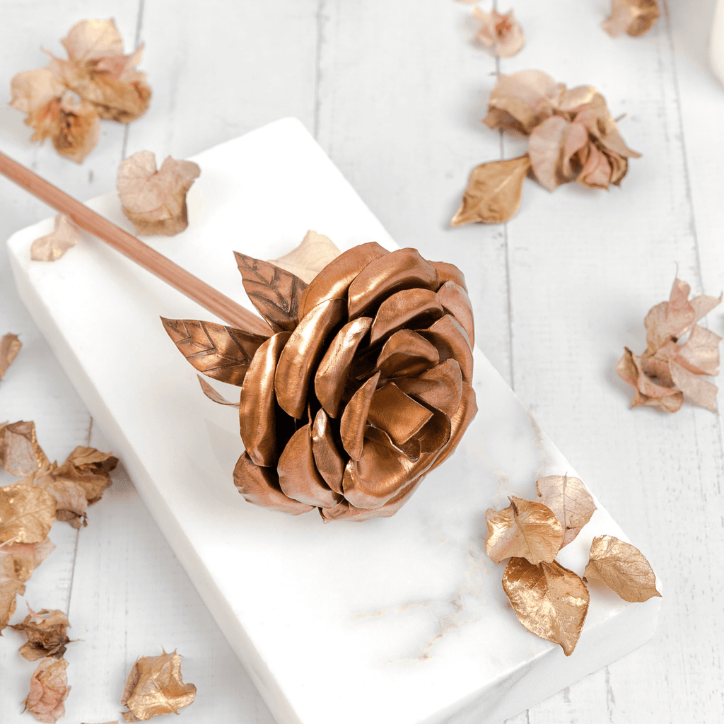 A rose ornament made entirely of copper, lays across a rectangular marble slab. Both items are atop a white, wooden base surrounded by copper leaves. The rose is made by Empire Copper.