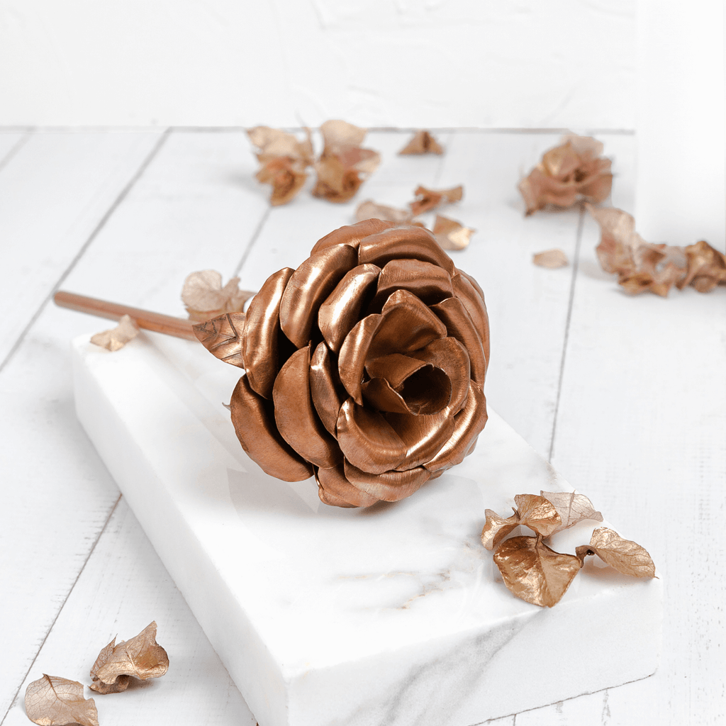 A rose ornament made entirely of copper, lays across a rectangular marble slab. Both items are atop a white, wooden base surrounded by copper leaves. The rose is made by Empire Copper.