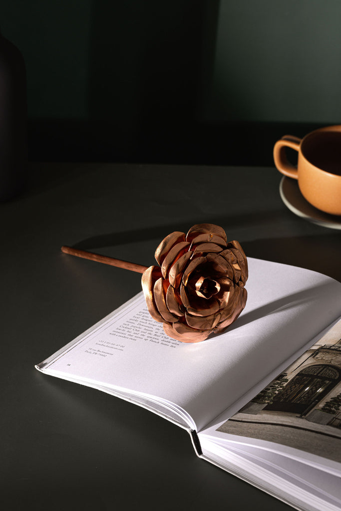 A rose made entirely from copper, laying across an open, white page book. In the background to the right, you can partially see a brown teacup and saucer. They items are sitting atop a moss green table. The copper rose is made by Empire Copper.