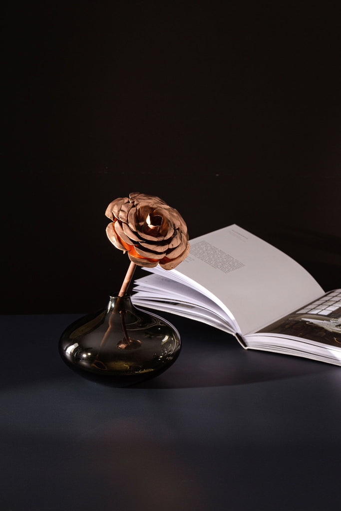 A single, long stem rose made entirely from copper. The rose is sitting up in a small glass jar. In the background, to the right is an open book. All the items are atop a navy blue surface. The rose is made by Empire Copper.