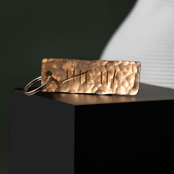 A keyring made from planished bronze and engraved with the eight tally. The keyring is balanced upright on top of a small, black box in front of a moss, green background. The keyring is made by Empire Copper.